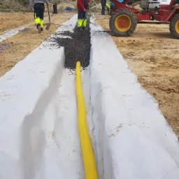 Wykop wyłożony białą geowłókniną, częściowo wypełniony żwirem, z żółtą rurą drenażową; w tle pracownicy z łopatami i wywrotka.