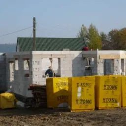 Wznoszenie ścian parterowego domu z bloczków Ytong, widoczne żółte opakowania materiału budowlanego, dwóch pracowników na budowie, w tle zielony dach innego budynku.