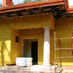 Żółta elewacja domu w trakcie ocieplania, widoczne białe kołki mocujące styropian, kolumna przy wejściu, częściowo odsłonięte drewniane elementy dachu.