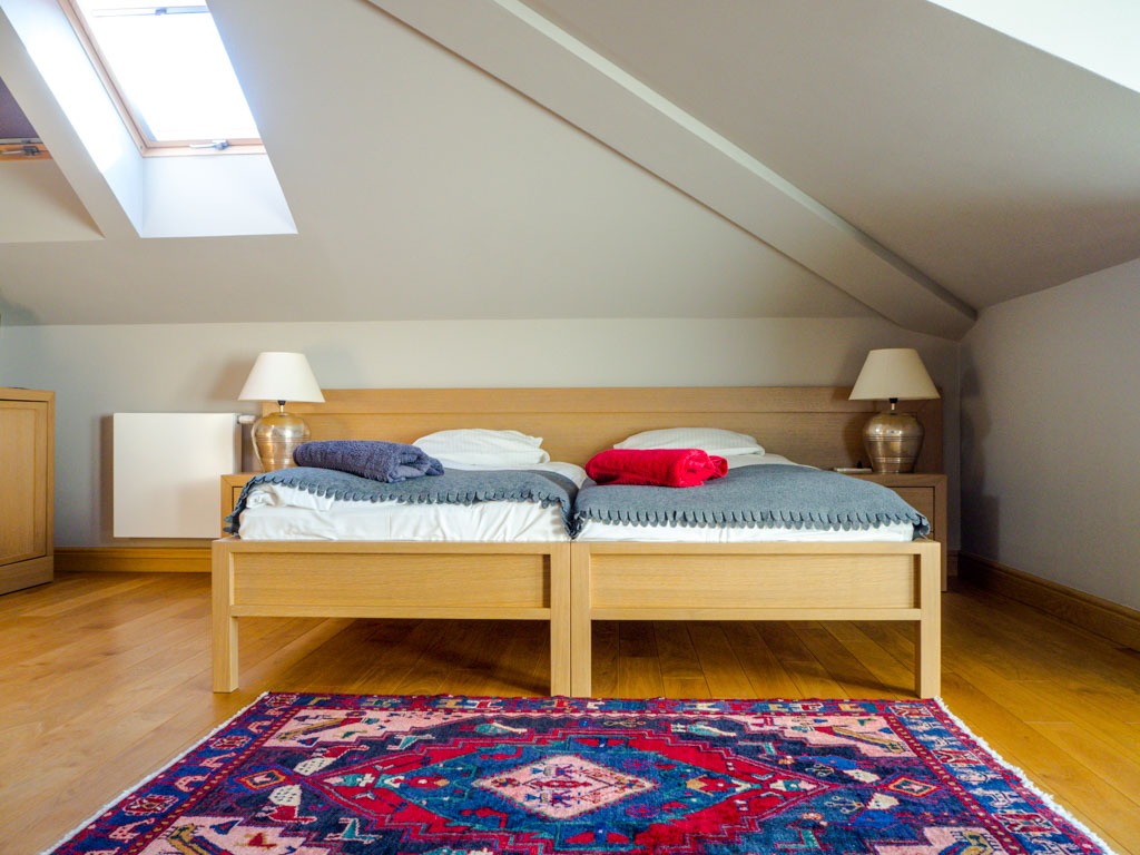 Pokój na poddaszu z dwoma pojedynczymi łóżkami z drewnianymi ramami, szarymi pledami, kolorowym dywanem i oknem dachowym.