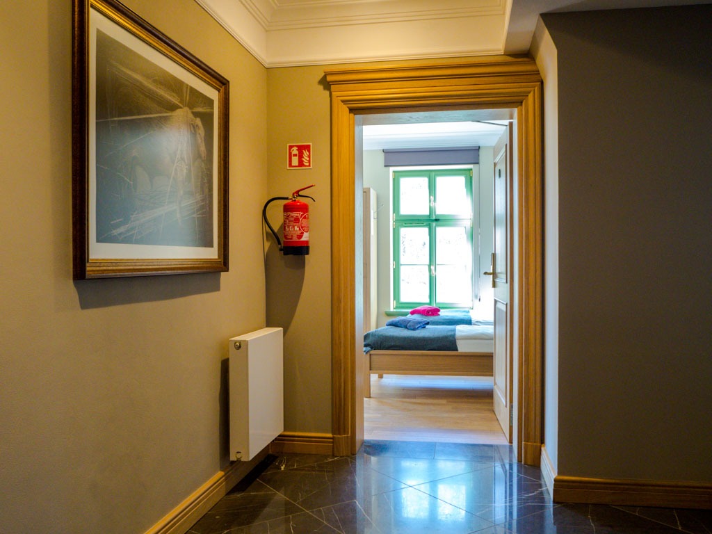 Korytarz z ciemną marmurową podłogą prowadzący do pokoju z łóżkami, na ścianie obraz w złotej ramie, gaśnica i biały grzejnik.