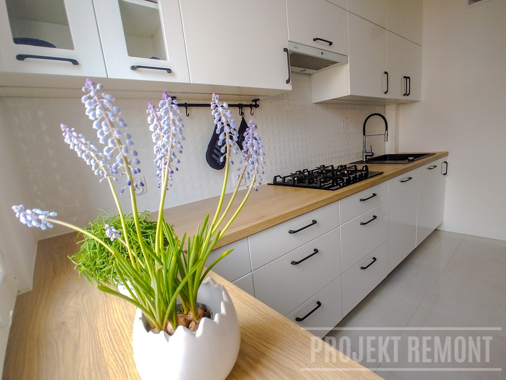 Nowoczesna, biała kuchnia z drewnianym blatem, czarnymi uchwytami i płytą gazową, ozdobiona bukietem szafirków w białej doniczce w kształcie jajka.