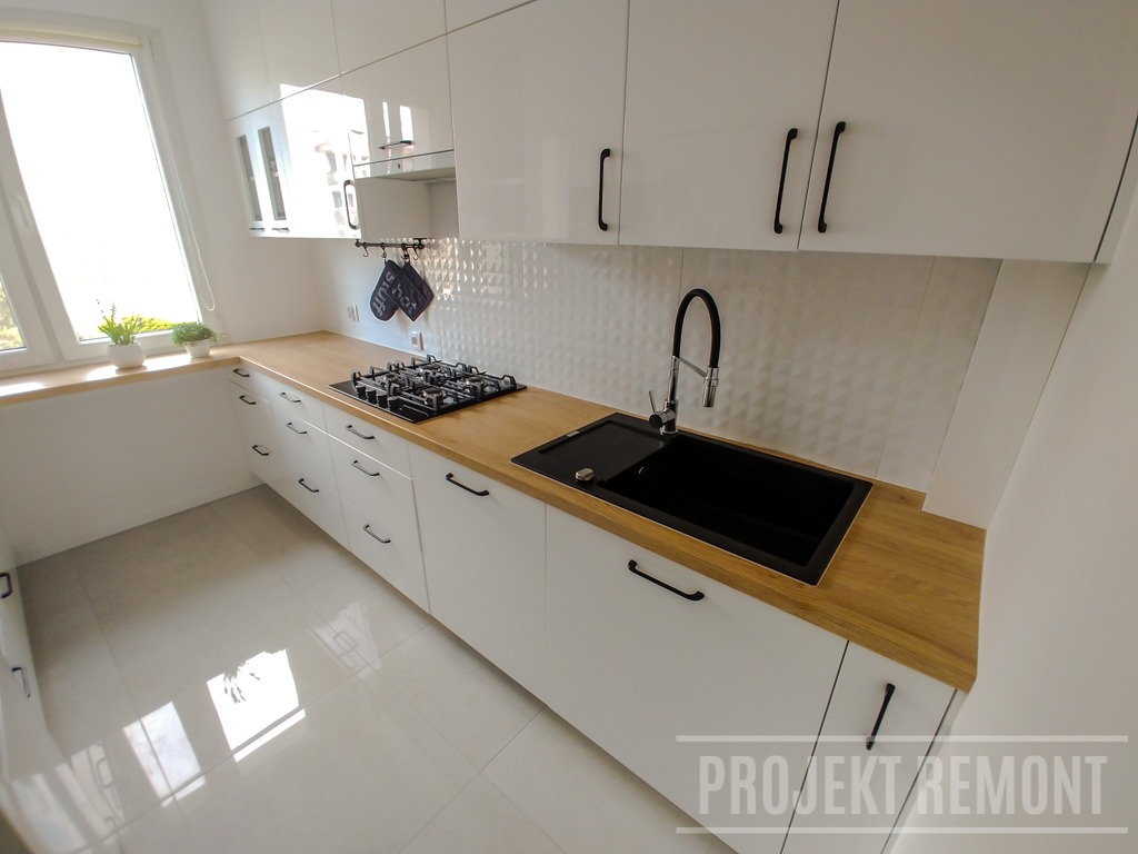 Nowoczesna, biała kuchnia z drewnianym blatem, czarnym zlewem i baterią, oraz płytą gazową. Na ścianie białe, trójwymiarowe płytki. Minimalistyczne czarne uchwyty szafek.