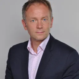 Portret mężczyzny w ciemnej marynarce i różowej koszuli, z niebieskimi oczami i lekko siwiejącymi włosami, na jasnoszarym tle.