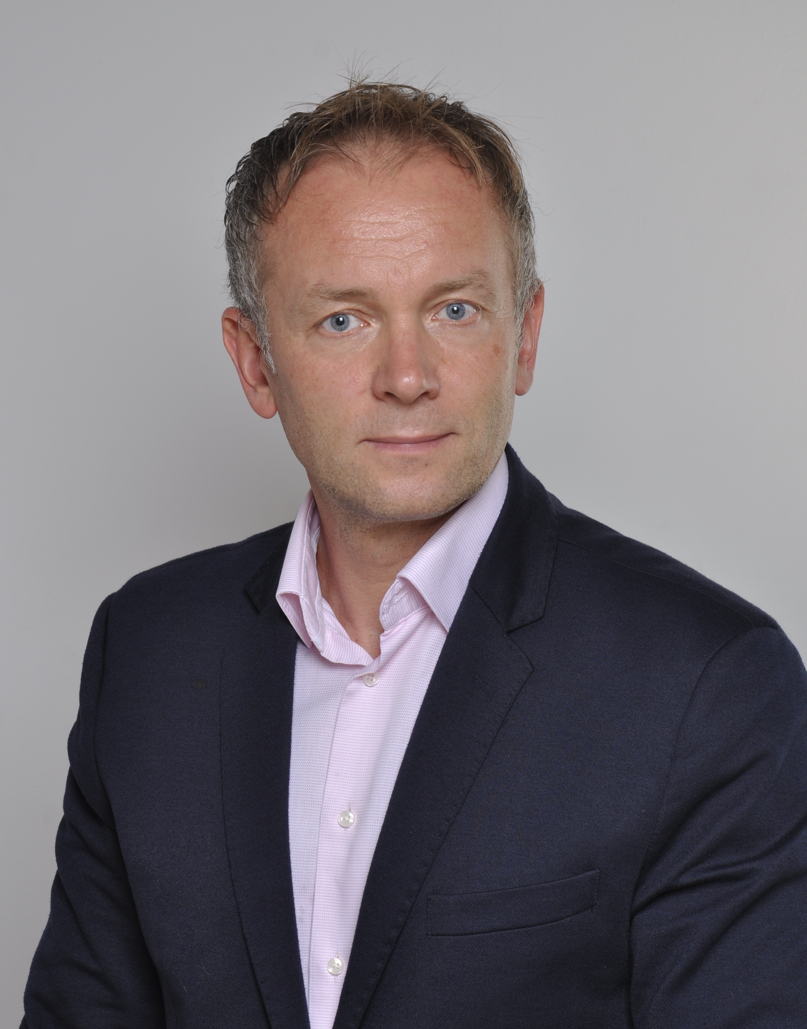 Portret mężczyzny w ciemnej marynarce i różowej koszuli, z niebieskimi oczami i lekko siwiejącymi włosami, na jasnoszarym tle.