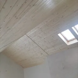 Poddasze z wykończoną drewnianą boazerią na skośnym suficie i oknem dachowym, ściany w stanie surowym przed malowaniem.