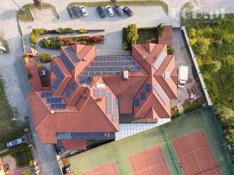 Widok z drona na rozległy dach z czerwonej dachówki, pokryty panelami słonecznymi, obok kort tenisowy i zaparkowane samochody.