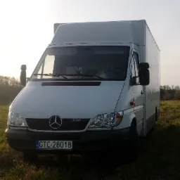 Biały furgon Mercedes-Benz 308 z zabudową kontenerową, zaparkowany na trawiastym polu w słoneczny dzień.