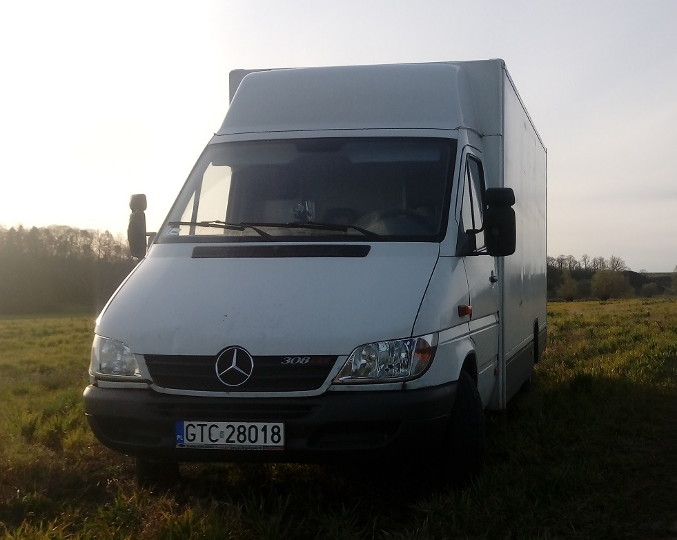 Biały furgon Mercedes-Benz 308 z zabudową kontenerową, zaparkowany na trawiastym polu w słoneczny dzień.