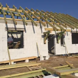 Budowa domu jednorodzinnego: wzniesione ściany z betonu komórkowego i konstrukcja dachu w trakcie montażu, widoczne drewniane elementy więźby dachowej i deski na ziemi.