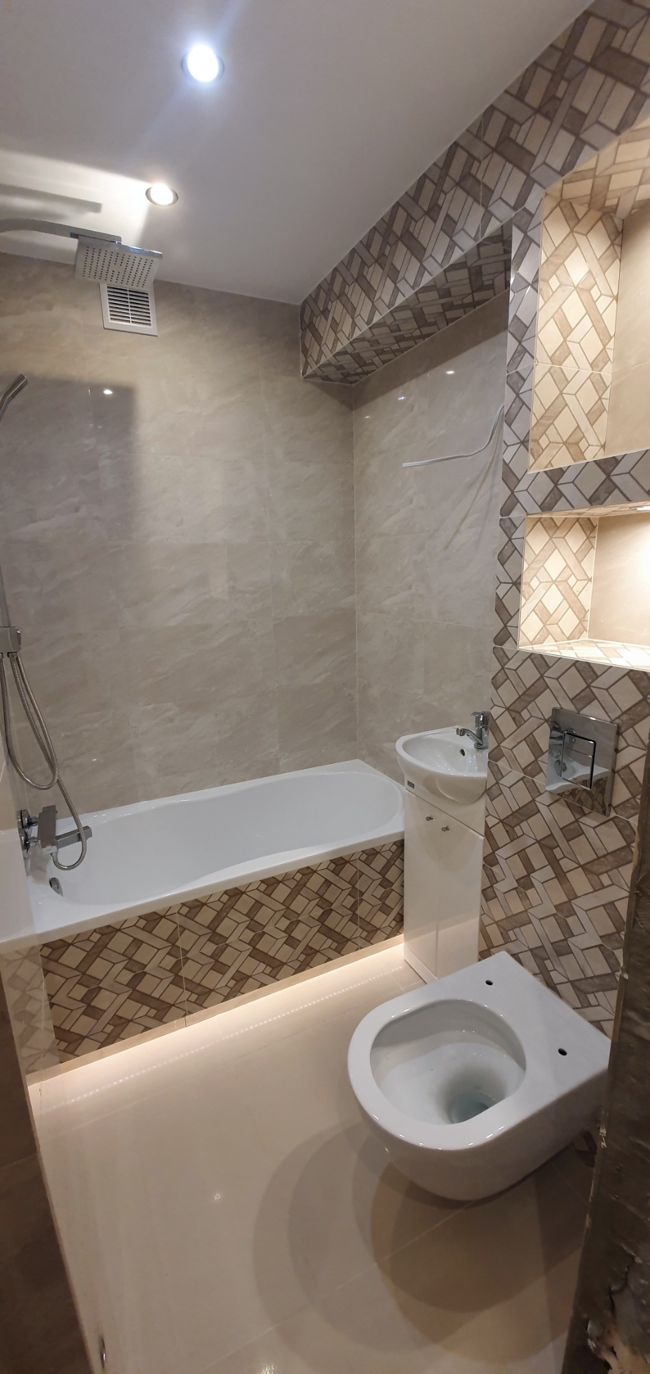 Wykończona łazienka z beżowymi płytkami imitującymi marmur, ozdobną mozaiką w geometryczne wzory na obudowie wanny i wnękach, podświetleniem LED pod wanną, białą armaturą: wanną, umywalką z szafką...