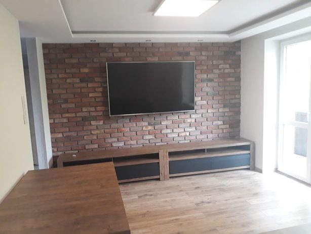 Salon z ceglaną ścianą, na której wisi telewizor, pod nim drewniana szafka RTV, podłoga wyłożona panelami imitującymi drewno.