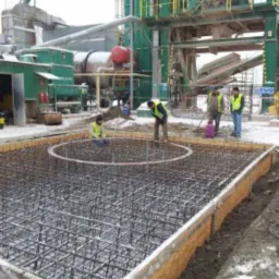 Przygotowanie zbrojenia pod fundament przemysłowy z widoczną konstrukcją stalową i pracownikami w kamizelkach odblaskowych na tle zakładu produkcyjnego.