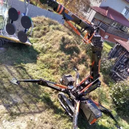 Mobilny mini dźwig gąsienicowy z przyssawkami próżniowymi podnosi szybę na tle domu jednorodzinnego i sterty drewna.