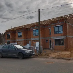 Budowa bliźniaka z zamontowanymi oknami i roletami zewnętrznymi w szarych ramach, widoczne drewniane elementy konstrukcji dachu, zaparkowany samochód osobowy na pierwszym planie.