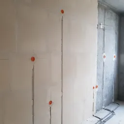 Ściana z płyt gipsowo-kartonowych z wyfrezowanymi bruzdami i puszkami instalacyjnymi pod gniazdka elektryczne, na ścianie betonowej widoczne poprowadzone kable w peszlach.