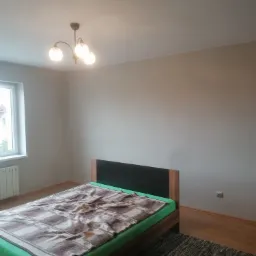 Pokój z łóżkiem i świeżo pomalowanymi ścianami w odcieniach beżu, widoczna lampa sufitowa i okno z radiatorem.