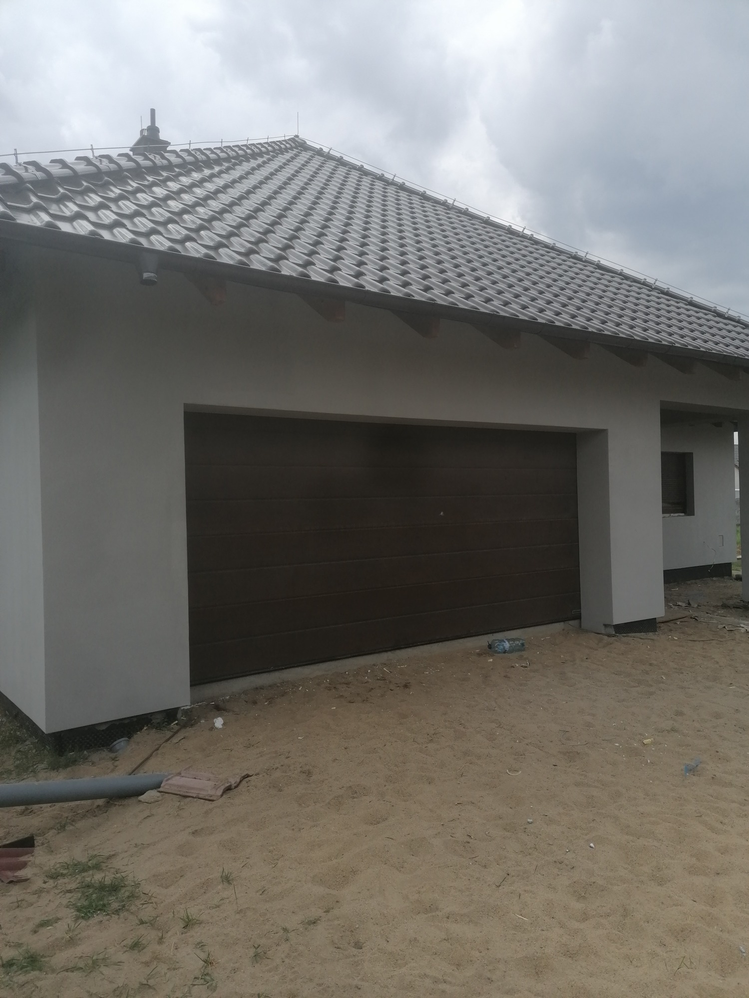 Świeżo otynkowany garaż z brązową bramą segmentową i szarą dachówką ceramiczną, widok zewnętrzny w pochmurny dzień, teren wokół jeszcze niezagospodarowany.