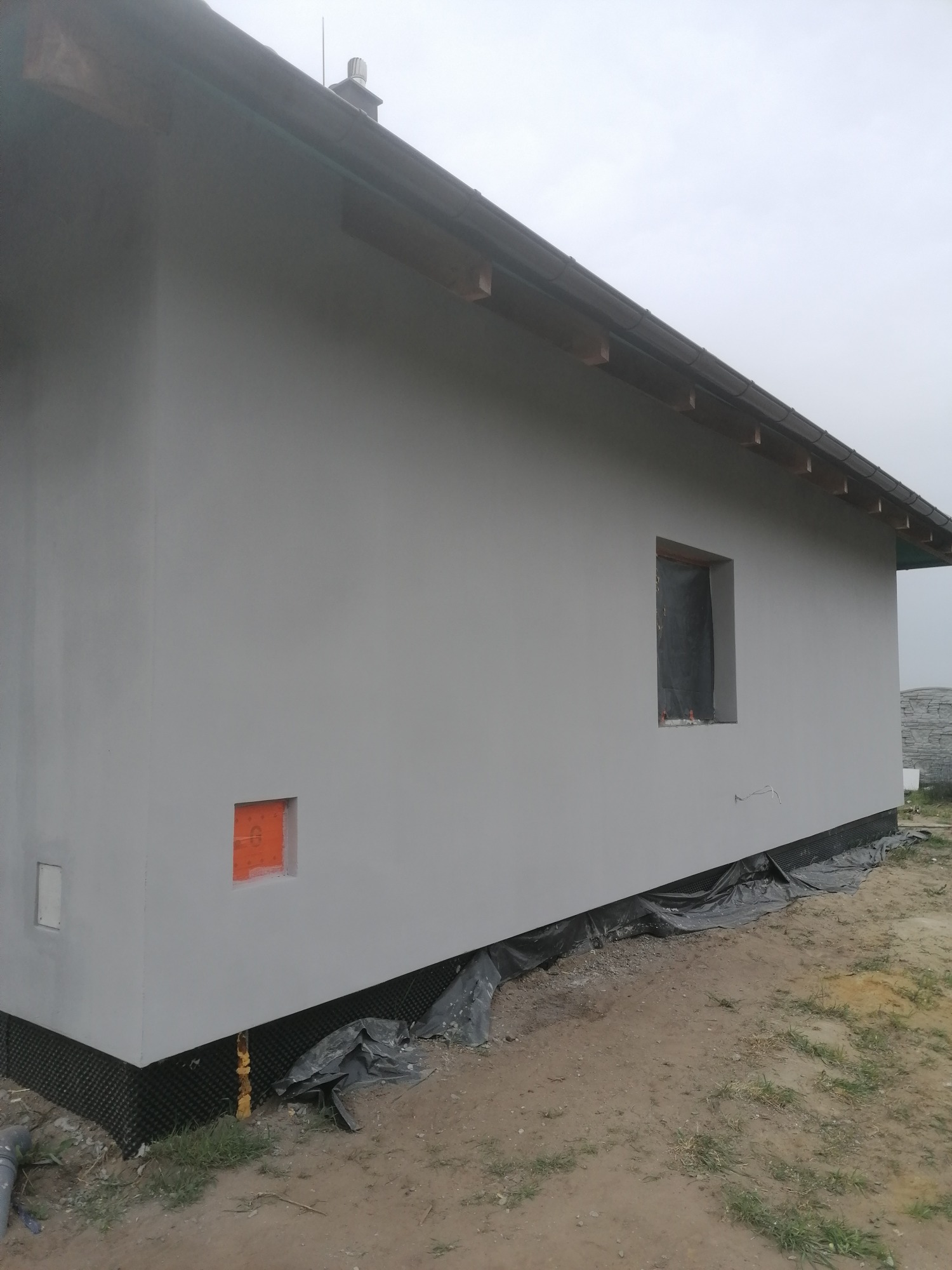 Świeżo otynkowana ściana zewnętrzna domu jednorodzinnego w kolorze szarym, z widocznymi otworami okiennymi zabezpieczonymi folią i fragmentem niezabezpieczonego otworu wentylacyjnego, ukazująca...