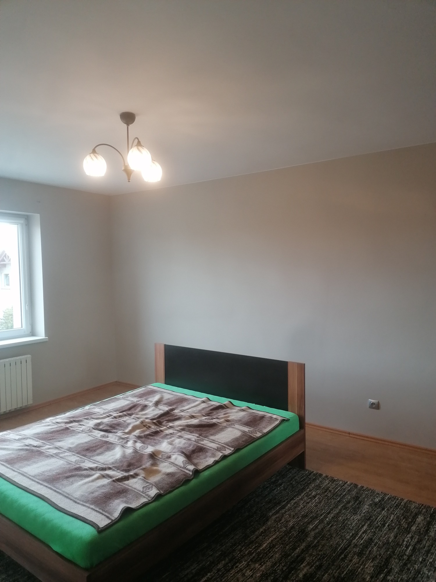 Pokój z łóżkiem i świeżo pomalowanymi ścianami w odcieniach beżu, widoczna lampa sufitowa i okno z radiatorem.