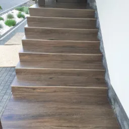 Schody zewnętrzne wyłożone brązowymi płytkami imitującymi drewno, z widocznymi detalami fug i krawędzi, prowadzące do drzwi wejściowych.