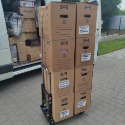 FHUP Sławomir Rączka - Wózek transportowy załadowany kartonami IKEA z napisanymi odręcznie nazwami zawartości, ustawiony przed otwartym busem wypełnionym dodatkowymi pudłami podczas dnia.