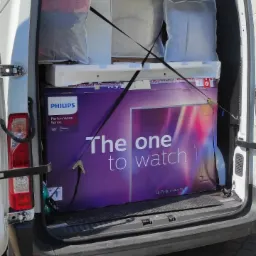 Wnętrze białego busa z otwartymi drzwiami bagażnika, załadowane kartonami i folią, zabezpieczone pasami transportowymi, w centrum duży karton z telewizorem Philips 'The one to watch'.