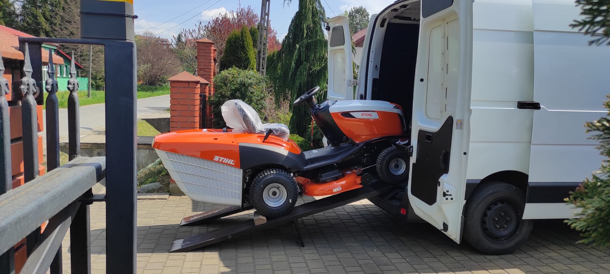 Pomarańczowo-biała kosiarka traktorek marki Stihl wjeżdża po rampie do białego busa dostawczego, widok z zewnątrz na posesję z ogrodzeniem.