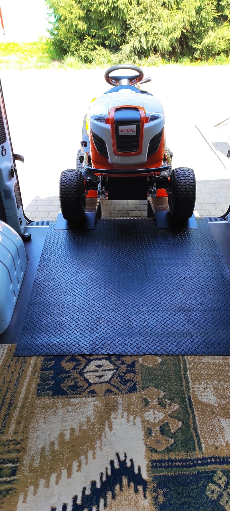 Traktorek do koszenia trawy marki Stihl przewożony w busie z rampą załadunkową, widok z tyłu pojazdu na urządzenie na tle zieleni.