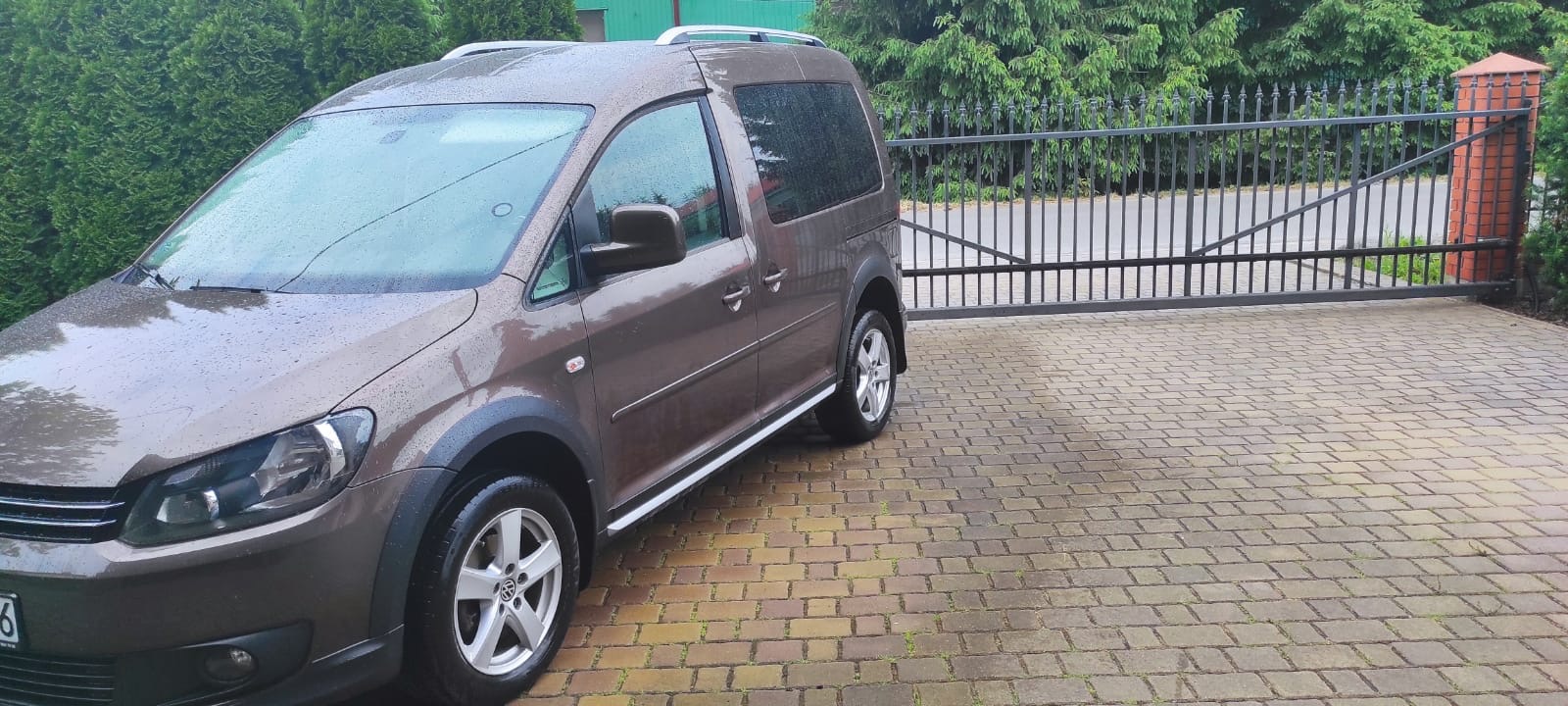 Brązowy Volkswagen Caddy z relingami dachowymi, zaparkowany na brukowanym podjeździe przed czarną bramą, widoczne krople deszczu na karoserii.