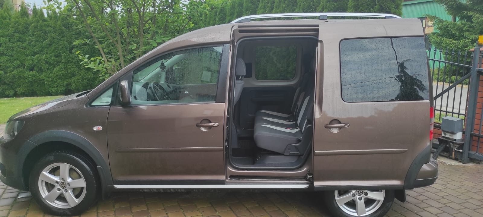 Brązowy Volkswagen Caddy z otwartymi przesuwnymi drzwiami, zaparkowany na brukowanej nawierzchni w deszczowy dzień, widoczne wnętrze z szarymi siedzeniami w paski.