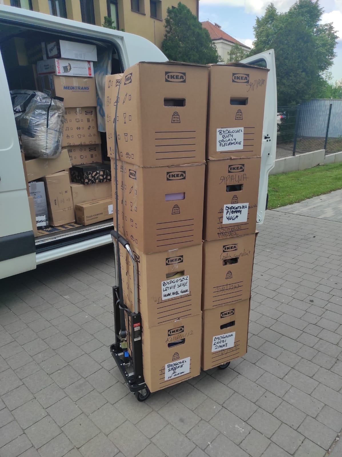 Wózek transportowy załadowany kartonami IKEA z napisanymi odręcznie nazwami zawartości, ustawiony przed otwartym busem wypełnionym dodatkowymi pudłami podczas dnia.