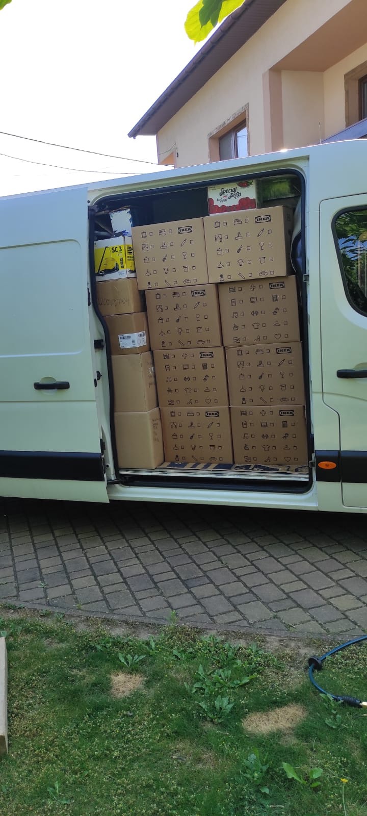 Wnętrze białego busa wypełnione po brzegi kartonami z logo IKEA, obok widoczny żółty odkurzacz i karton z pizzą, w tle dom jednorodzinny