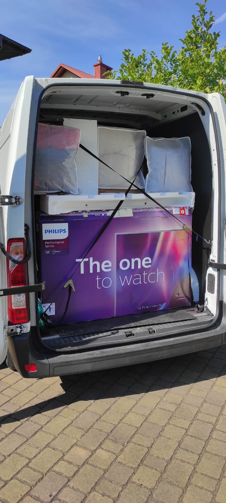 Wnętrze białego busa z otwartymi drzwiami bagażnika, załadowane kartonami i folią, zabezpieczone pasami transportowymi, w centrum duży karton z telewizorem Philips 'The one to watch'.