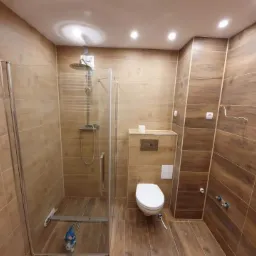 Wykończona łazienka z kabiną prysznicową ze szklanymi drzwiami, toaletą podwieszaną i brązowymi płytkami imitującymi drewno na ścianach i podłodze, widoczne niedokończone przyłącze elektryczne