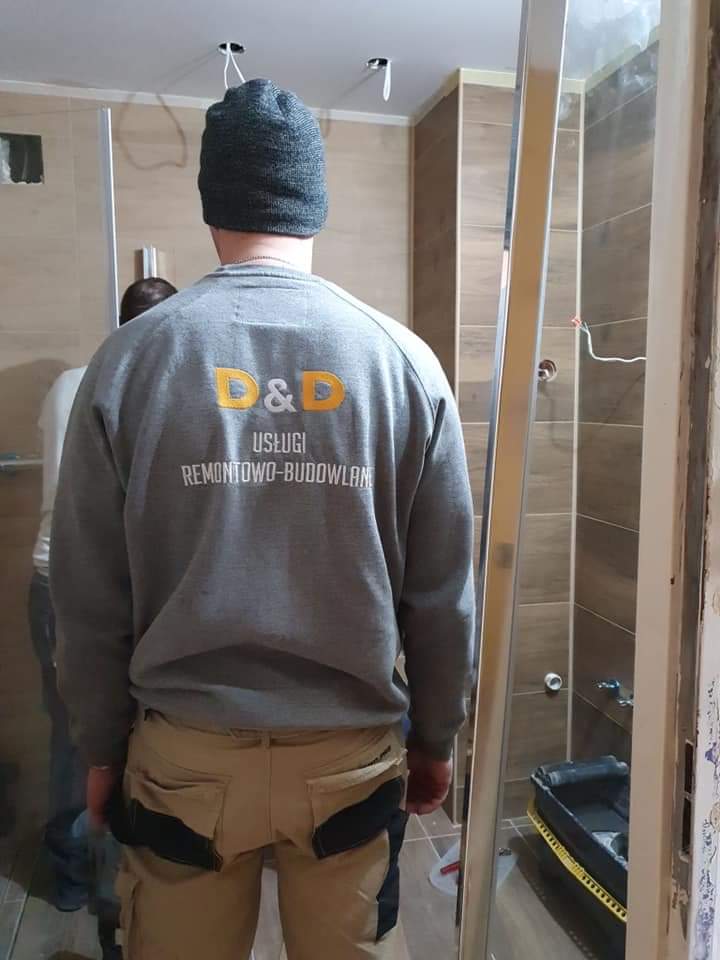 Mężczyzna w szarej bluzie z logo firmy remontowo-budowlanej, w trakcie prac w łazience z częściowo zamontowaną kabiną prysznicową i brązowymi płytkami na ścianach.