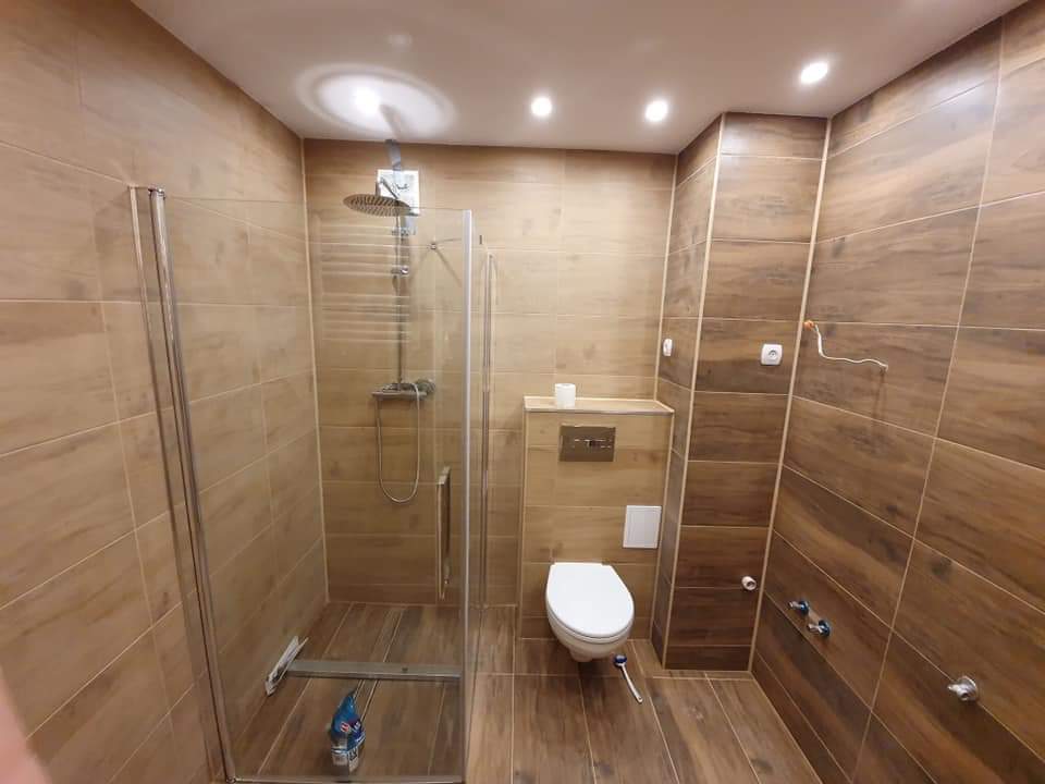 Wykończona łazienka z kabiną prysznicową ze szklanymi drzwiami, toaletą podwieszaną i brązowymi płytkami imitującymi drewno na ścianach i podłodze, widoczne niedokończone przyłącze elektryczne