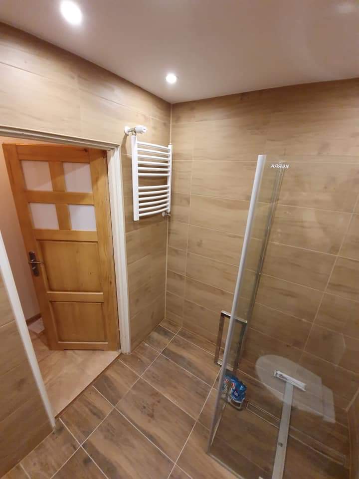 Wykończona łazienka z beżowymi płytkami imitującymi drewno, przeszklona kabina prysznicowa z widocznymi środkami czystości w środku, drewniane drzwi z czterema szybkami i grzejnik drabinkowy...