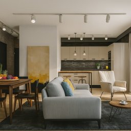 Sowa Design Projekty Wnętrz - Jasne, przestronne wnętrze kawalerki z ceglaną ścianą, drewnianym stołem i szarą sofą. Widoczna kuchnia w odcieniach beżu i czerni. Całość w nowoczesnym stylu.