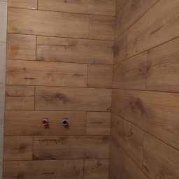 Daniel Brychcy - Ściana wyłożona brązowymi płytkami imitującymi drewno, widoczne dwa wystające przyłącza wodne z zaślepkami w kolorze czerwonym i niebieskim.