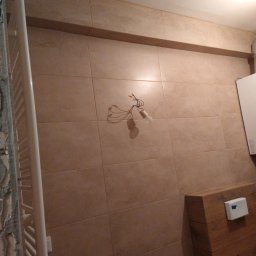 Daniel Brychcy - Łazienka w trakcie prac: beżowe płytki ścienne, grzejnik drabinkowy z resztkami zaprawy, drewniana obudowa toalety, wisząca żarówka z kablami.