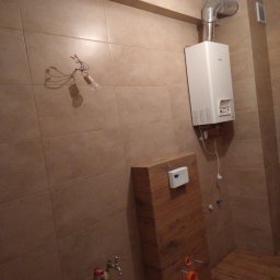 Daniel Brychcy - Remontowana łazienka z beżowymi płytkami, wystającymi kablami z żarówką, częściowo zamaskowaną instalacją podtynkową WC obudowaną drewnem i zamontowanym piecykiem gazowym.