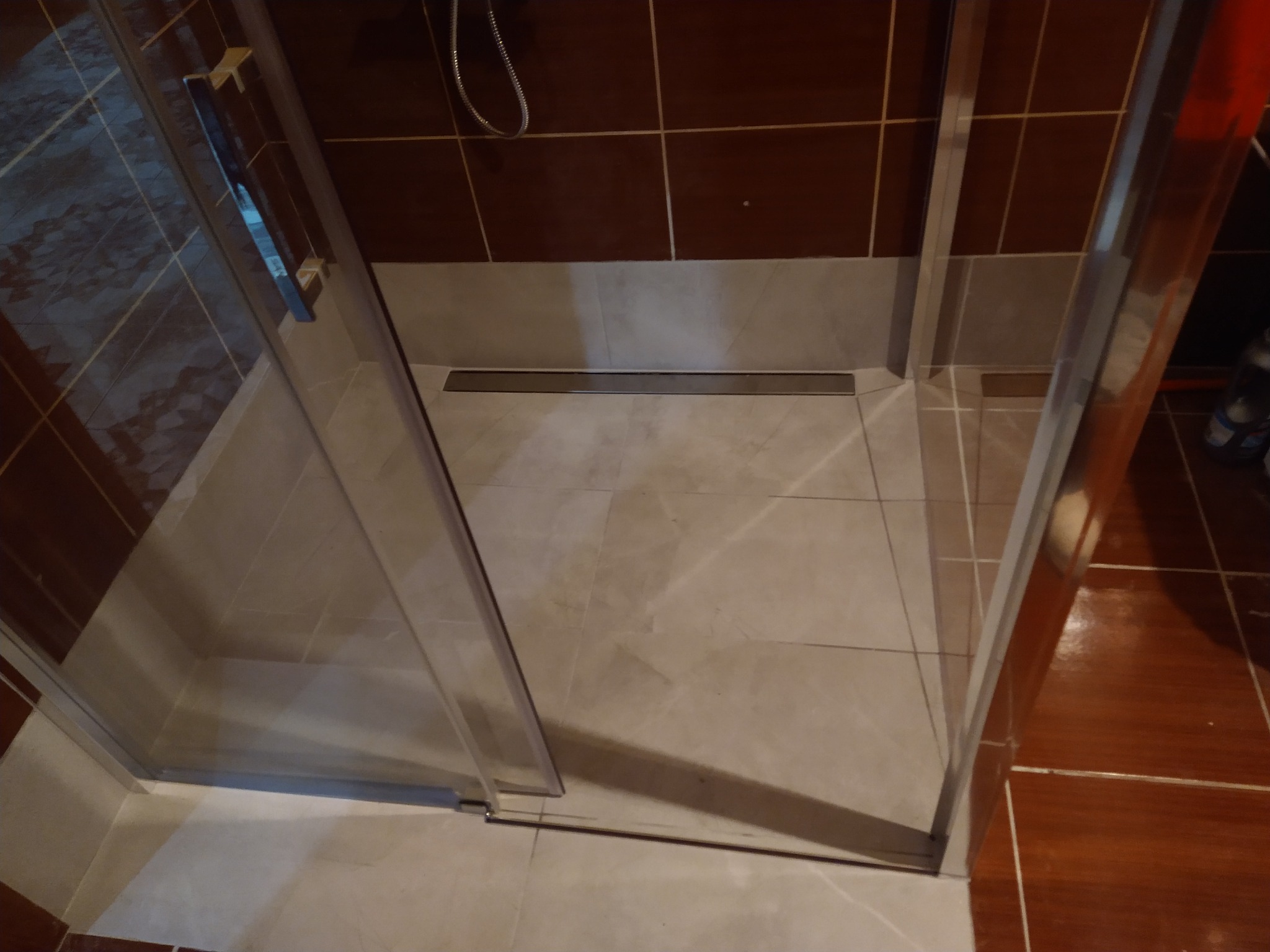 Kabina prysznicowa z odpływem liniowym, wykończona brązowymi i beżowymi płytkami, widoczne detale uszczelnień i mocowań szkła.