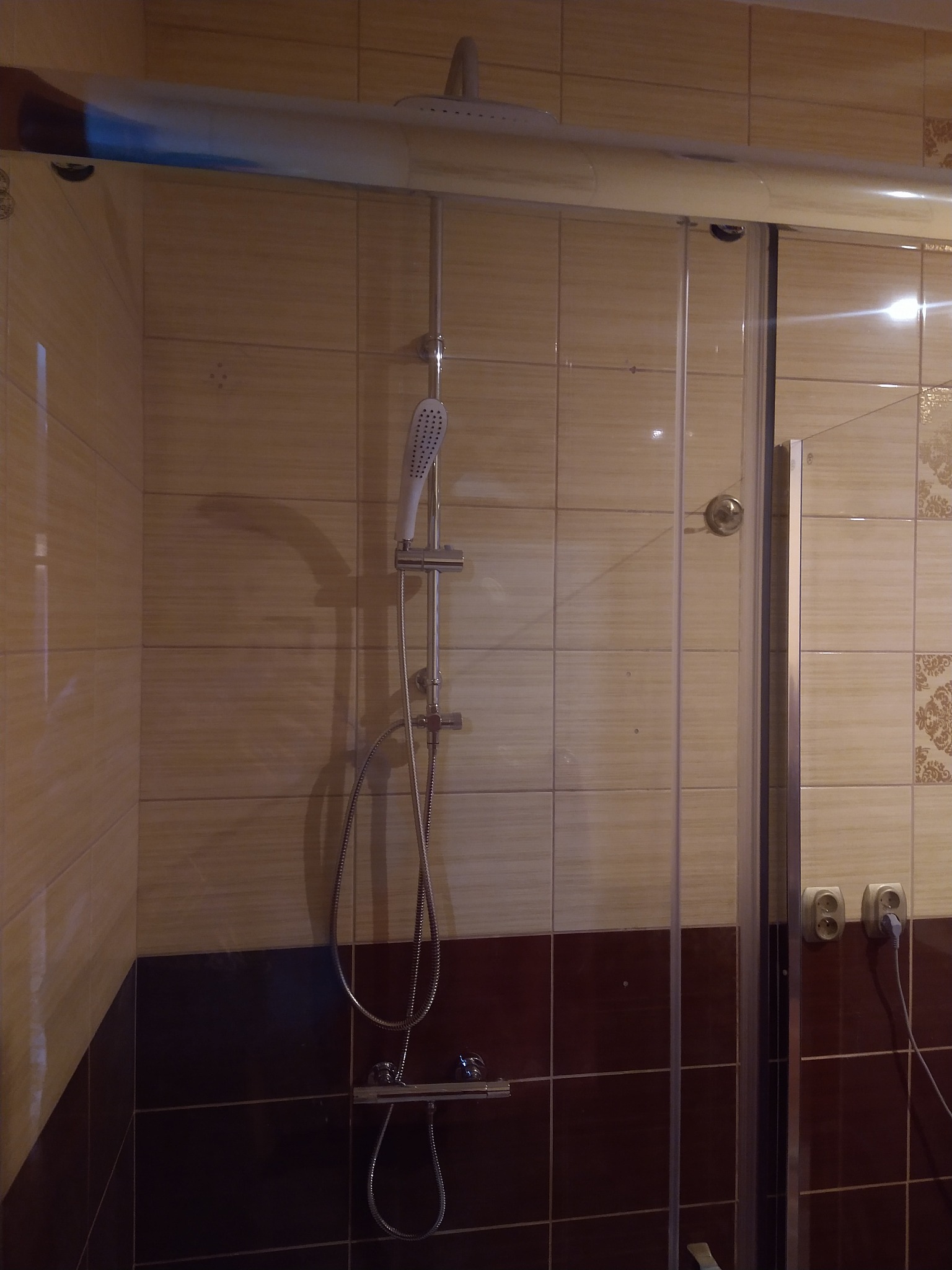 Kabina prysznicowa z przesuwnymi drzwiami, widoczne beżowe i brązowe płytki na ścianach, biała słuchawka prysznicowa na regulowanym drążku i bateria termostatyczna.