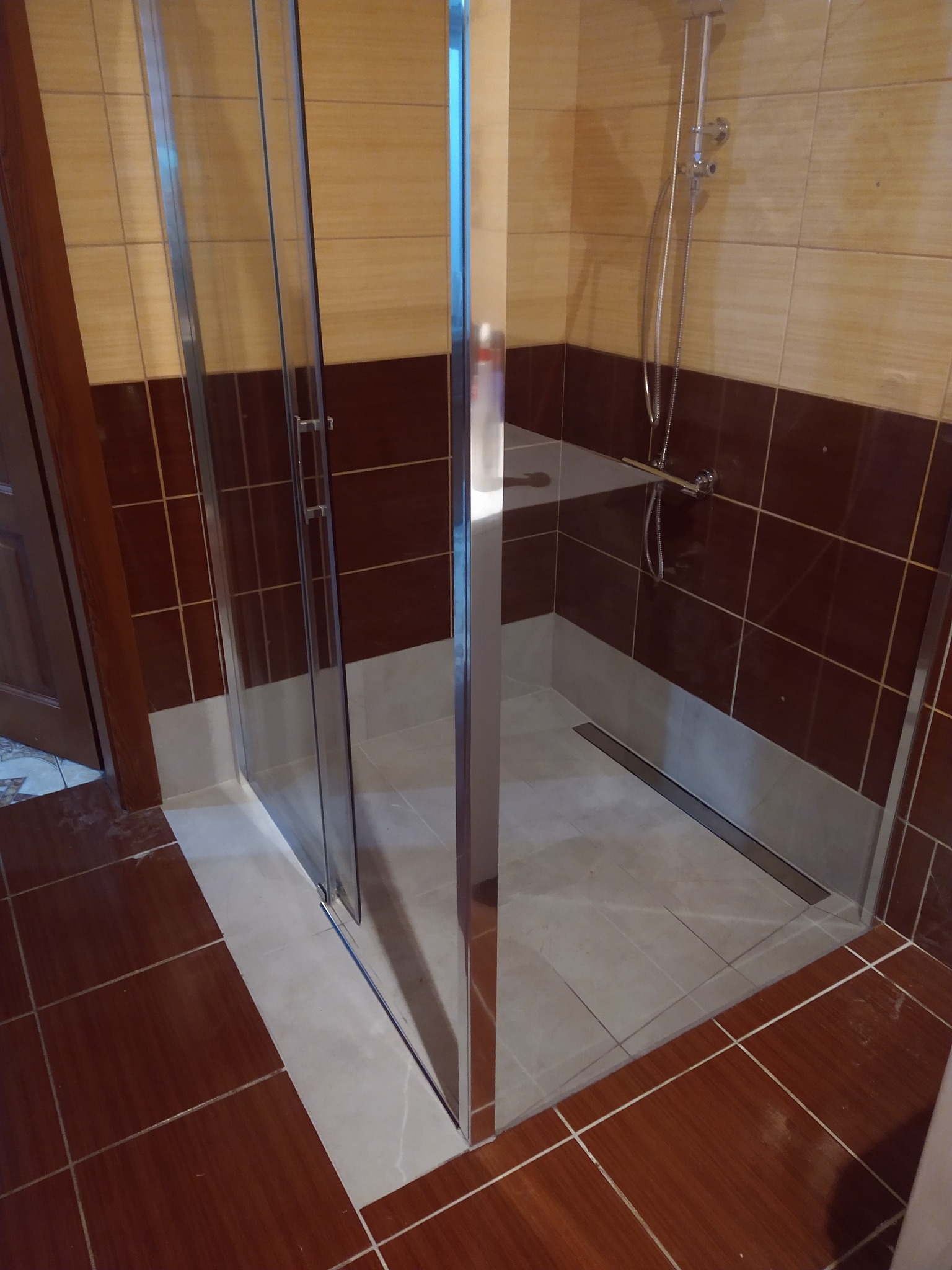 Kabina prysznicowa z chromowanymi profilami i odpływem liniowym, wykończona brązowo-beżowymi płytkami ceramicznymi z widoczną armaturą natryskową.