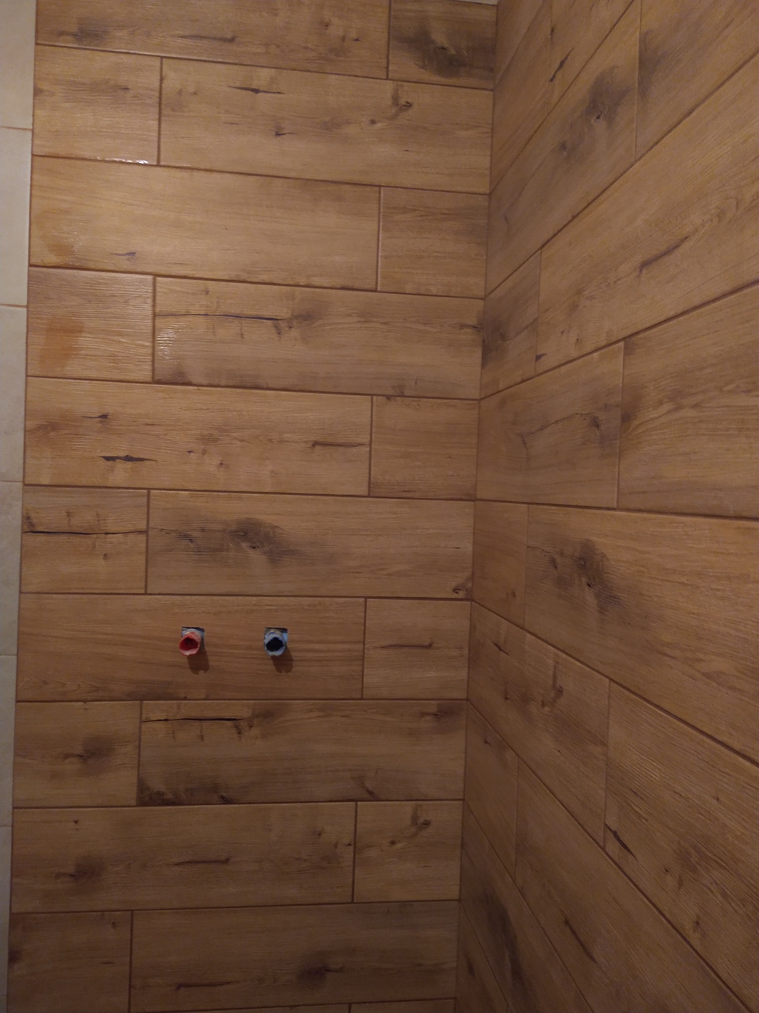 Ściana wyłożona brązowymi płytkami imitującymi drewno, widoczne dwa wystające przyłącza wodne z zaślepkami w kolorze czerwonym i niebieskim.