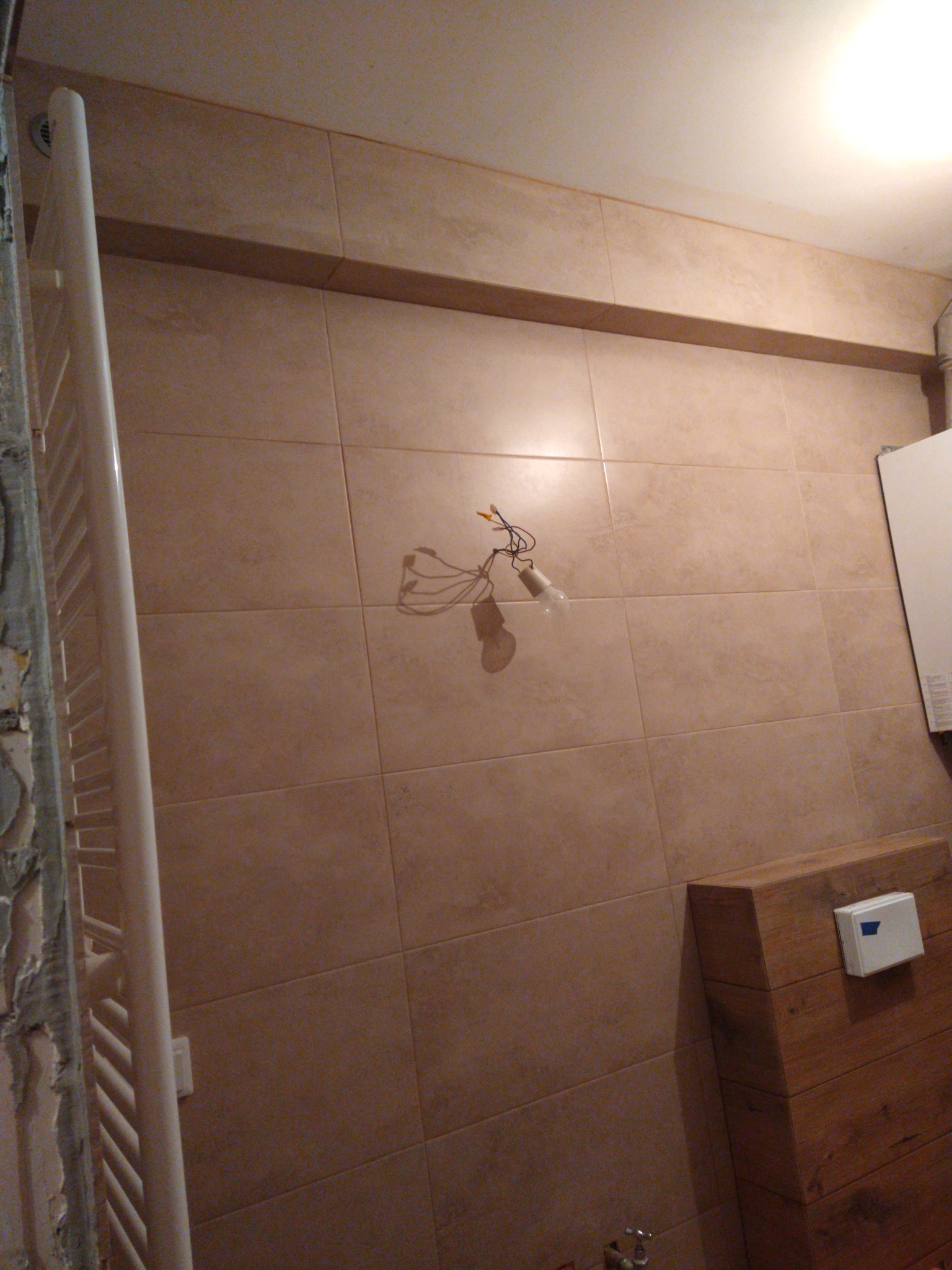 Łazienka w trakcie prac: beżowe płytki ścienne, grzejnik drabinkowy z resztkami zaprawy, drewniana obudowa toalety, wisząca żarówka z kablami.