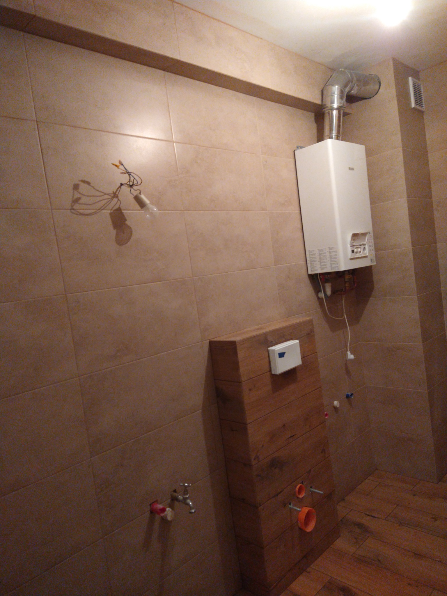 Remontowana łazienka z beżowymi płytkami, wystającymi kablami z żarówką, częściowo zamaskowaną instalacją podtynkową WC obudowaną drewnem i zamontowanym piecykiem gazowym.