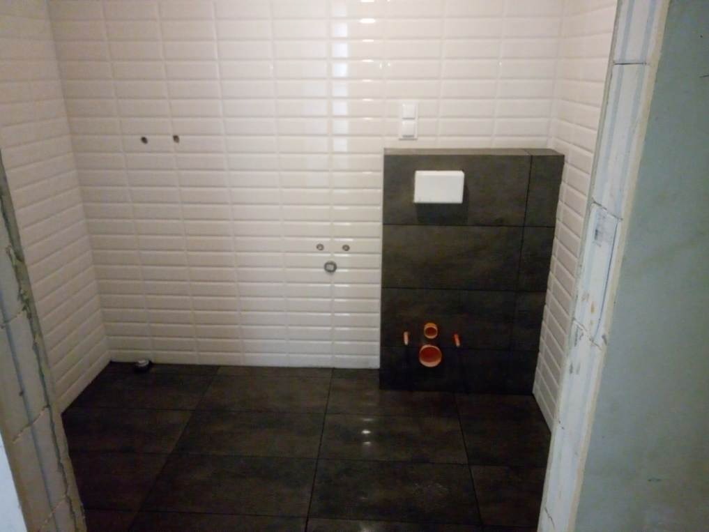 Łazienka w trakcie remontu: ułożone ciemne płytki podłogowe, białe płytki na ścianach, zabudowa stelaża WC z szarych płytek, widoczne rury i instalacje.