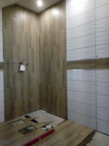 Układanie płytek imitujących drewno na podłodze i ścianach w łazience, widoczny poziomica i fragment instalacji hydraulicznej w podłodze, białe płytki faliste na ścianie po prawej.